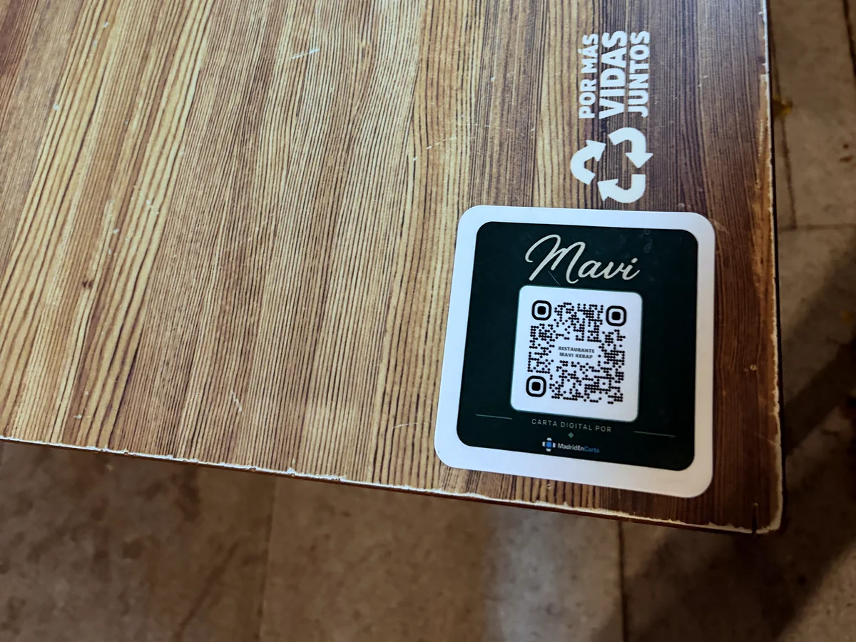 Esquina de una mesa de la terraza de Restaurante Mavi Kebap con el vinilo verde de la carta digital pegado: el nombre Mavi, el QR y «Carta digital por MadridEnCarta»